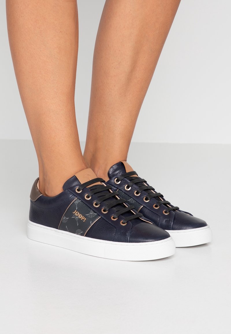 Baskets en cuir bleu marine avec un panneau latéral à motif, des accents beige et une semelle en caoutchouc blanche. Elles présentent des lacets foncés et une languette avec logo.