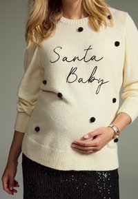 Cream-colored sweater with black pom-poms, featuring the text "Santa Baby" in cursive. Worn over a black sequined skirt.