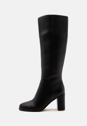 Botte en cuir noire montant jusqu’au genou avec un bout arrondi et un talon épais et haut, vue de profil sur un fond clair uni.