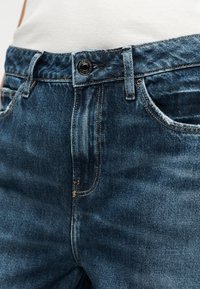 I jeans di denim blu presentano una cintura aderente con un bottone metallico, cuciture a contrasto e un motivo sbiadito sottile su tutto il tessuto.