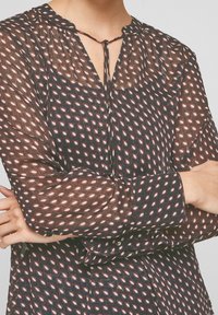 Blusa trasparente e scura con bottoni, scollatura a V e dettaglio con fiocco. Decorata con forme a diamante alternate in rosso e bianco su uno sfondo blu navy.