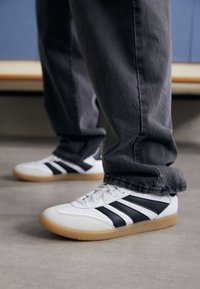 Des chaussures en cuir blanc avec des rayures noires, une semelle en caoutchouc gomme, un bout arrondi et un design bas, portées avec un pantalon en denim gris.