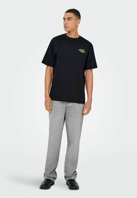 Sort bomulds T-shirt med rund halsudskæring og korte ærmer, med en grøn tekstdesign. Paarret med lysegrå lige ben jeans og sorte sko.