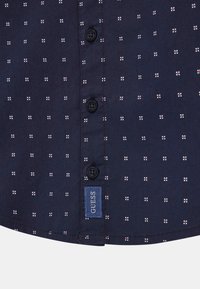 Camicia blu navy con un piccolo motivo a pois bianchi e rossi, dotata di bottoni neri e di un'etichetta blu "GUESS" in basso a sinistra.