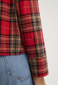 Camicia a quadri rossa e beige con maniche lunghe, caratterizzata da dettagli neri e un tessuto testurizzato, abbinata a jeans in denim blu chiaro.