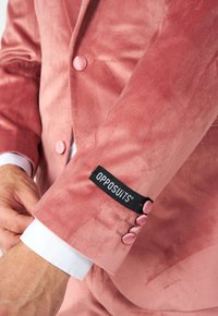 Chaqueta de traje de terciopelo rosa con un brillo sutil, botones redondeados y una etiqueta negra con "OPPOSUITS". Puño de camisa blanca visible por debajo.