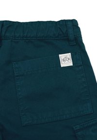 Tumma tiilenvärinen puuvillahousu, jossa on joustovyötärö, neliömuotoinen takatasku ja kontrastivärinen ommeltu logotagi.