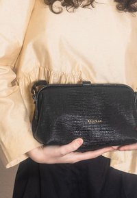 Černá texturovaná kožená kabelka přes rameno se zlatým logem, obdélníkového tvaru a s malým popruhem, držená na světlé blůze.