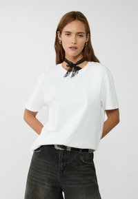 T-shirt bianca in cotone con maniche corte e scollatura rotonda, abbinata a jeans scuri e un choker in pizzo nero legato a forma di fiocco.
