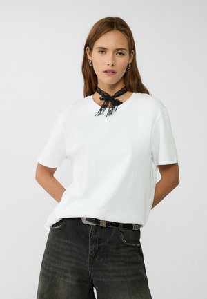 T-shirt bianca in cotone con maniche corte e scollatura rotonda, abbinata a jeans scuri e un choker in pizzo nero legato a forma di fiocco.