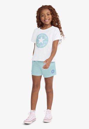 Niña sonriente con cabello rizado que lleva una camiseta blanca de Converse All Star, pantalones cortos azul claro con logo de estrella, y zapatillas blancas de caña alta, posando sobre fondo blanco.