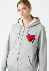 Felpa grigia con zip e cappuccio foderato in crema, caratterizzata da un cuore ricamato sul petto e dettagli con spille di sicurezza in nero.