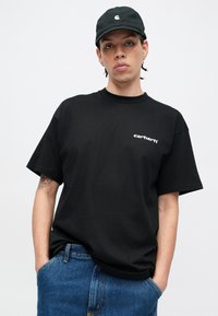 Camiseta de algodón negra con mangas cortas, con el logo blanco de Carhartt en el pecho. Combinada con pantalones cortos de mezclilla azul, ajuste casual.