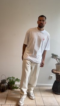 T-shirt bianca oversize con grafica rossa, pantaloni larghi color crema e sneaker bianche con dettagli neri. Ambientazione interna con piante.