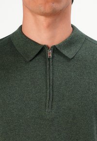 Artimiausias žmogaus kaklo ir viršutinės krūtinės vaizdas, vilkintis žalią megztą polo marškinėlį su užtrauktuku, kuris yra dalinai užtrauktas.