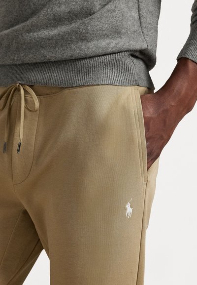 Beige sporta bikses, kas izgatavotas no mīkstas auduma, ar sašaurināmu jostasvietu un sānu kabatām. Izšūts balts logo uz kreisā kājas.