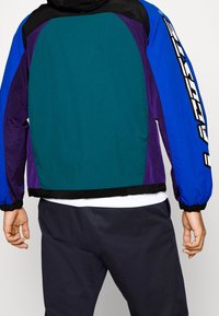 Color-blocked jacket featuring teal, purple, and blue panels. Lightweight, with elastic cuffs and a prominent logo down the sleeve.