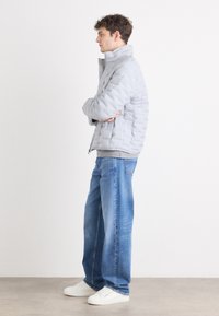 Lichtgrijze pufferjack met een gestikte textuur, hoge kraag en ritsluiting. Draagt over een grijze trui, gecombineerd met een blauwe spijkerbroek en witte sneakers.