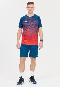 Man som står, iklädd en sporttröja med röd och blå mönster, blå shorts, vita strumpor och svarta sportskor framför en vit bakgrund.