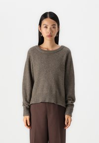 Maglione a maglia marrone castano con maniche lunghe, scollatura ampia e orlo a coste. Abbinato a pantaloni marroni, su uno sfondo neutro.