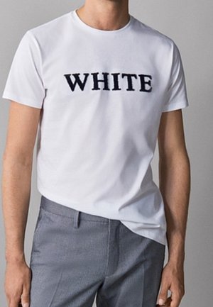White cotton t-shirt featuring bold black lettering "WHITE" across the chest. Short sleeves, crew neck; worn with gray trousers.