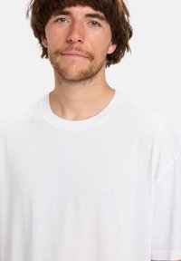 Jonge man met een lichte baard en bruin haar in een eenvoudige witte t-shirt met ronde hals tegen een witte achtergrond.
