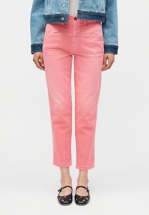 Γυναίκα φοράει κοντά ροζ τζιν παντελόνια συνδυασμένα με μαύρα παπούτσια Mary Jane με τρουκς και ένα μπλε τζιν μπουφάν, στέκεται μπροστά σε λευκό φόντο.