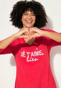 Punainen V-aukkoa oleva paita, jossa on kolme neljännesosaa hihat ja valkoiset tekstit "JE T'AIME bien" sekä tähtikoristeet. Pehmeä ja sileä kangas, rento istuvuus.