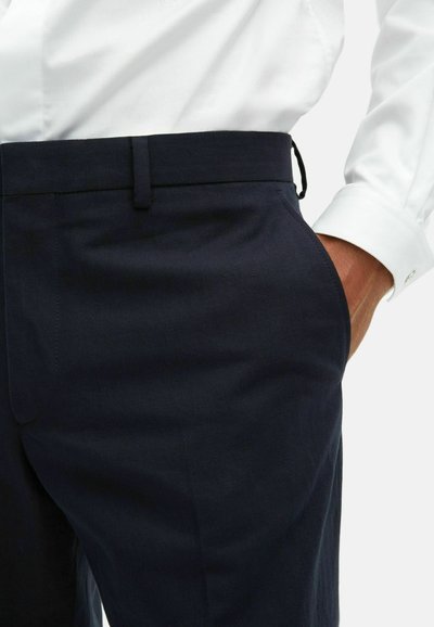 Mężczyzna w granatowych spodniach i białej koszuli z ręką w kieszeni na białym tle.