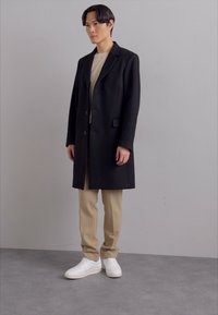 Svart ullkappa med en strukturerad design, knappstängning och två fickor, bärs över en beige topp och beige byxor. Vita sneakers fulländar looken.