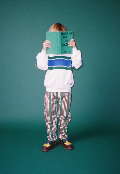 Maglione bianco con strisce blu e verdi, pantaloni a righe multicolori e scarpe chunky. Tiene un libro verde intitolato "REGOLAMENTI INVENTATI DA ME."