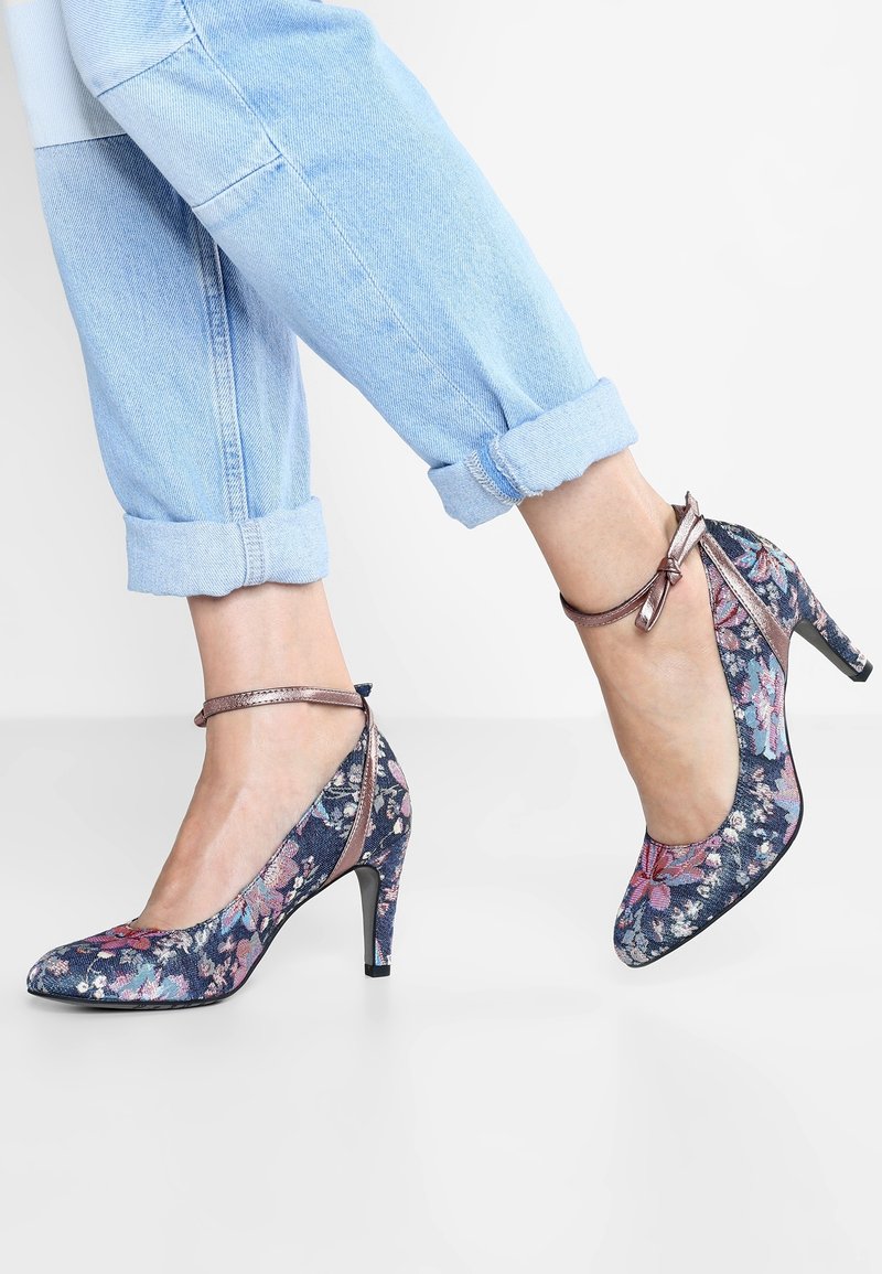 Jambes en jean bleu clair retroussé portant des talons hauts bleus à fleurs avec des brides à la cheville et des nœuds sur fond blanc.