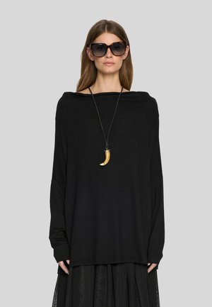 Kobieta z długimi brązowymi włosami, nosząca przerośnięte czarne okulary przeciwsłoneczne, luźną czarną bluzkę, długą czarną spódnicę oraz naszyjnik z dużym zawieszką w kształcie rogu.