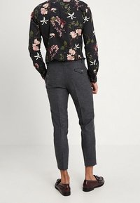 Zwarte bloemenshirt met roze en groene accenten, gecombineerd met grijze tapered broek en donkere loafers. Opvallende details zijn de contrasterende texturen.