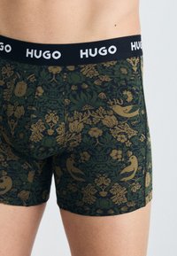 Grüne gemusterte Boxershorts mit einem Blumen- und Vogeldesign, einer schwarzen elastischen Taille mit dem Markennamen "HUGO" in Weiß.