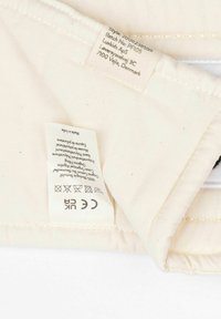 Kermanvärinen kangas, jossa hoito-ohjeita ja valmistajan tiedot sisältävät tarrat, ommellut reunat ja sileä pinta.