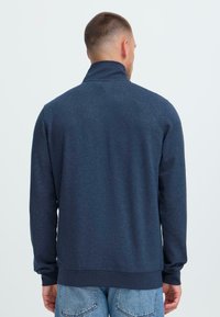 Muž s krátkymi vlasmi, ktorý má na sebe tmavomodrý pulóver s dlhým rukávom a svetlomodré džínsy, zobrazený zozadu na jednofarebnom pozadí.