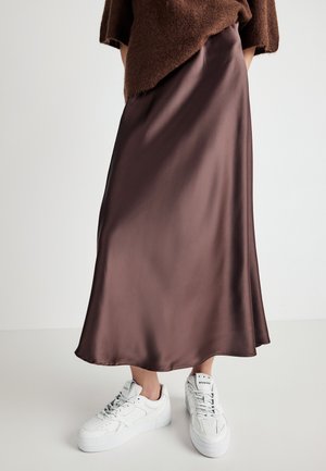 Person in einem braunen Satin-Midirock mit asymmetrischem Saum und weißen Plateau-Sneakern vor einem einfarbigen Hintergrund.