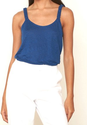 Kobieta ubrana w niebieską bluzkę bez rękawów z marszczonymi ramiączkami oraz białe spodnie z wysokim stanem, stojąca na neutralnym tle.