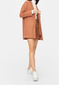 Frau in einem weißen Rollkragenpullover, brauem knopfartigen Rock, rostfarbenem Mantel und weißen Turnschuhen, die vor einem schlichten hellen Hintergrund steht.