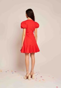 Sarkans īss kleita ar pūkainām piedurknēm, platu apakšmalu un teksturētu audumu. Apvienota ar bēša augstpapēžu sandālēm; uz zemes izkaisīta konfeti.