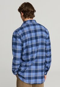 Uomo di spalle che indossa una camicia a maniche lunghe blu e blu scuro a quadri e pantaloni marroni su uno sfondo neutro.