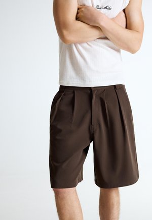 Man wearing dark brown pleated shorts and a white sleeveless top with arms crossed against a plain light background.