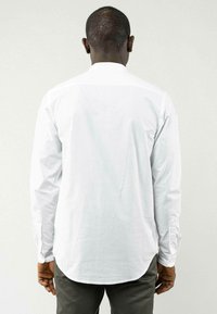 Weißes Langarmhemd mit leicht abgerundetem Saum. Baumwollstoff mit geknöpften Manschetten. Rückansicht zeigt klare Linien und minimales Design.