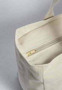 Plátěná taška v krémové barvě, s uzávěrem na zip se zlatými detaily a strukturovaným, obdélníkovým tvarem se šitými okraji.