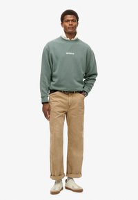 Sweatshirt in muted green with a high collar, featuring a white logo. Paired with rolled beige pants and cream sneakers with brown soles.