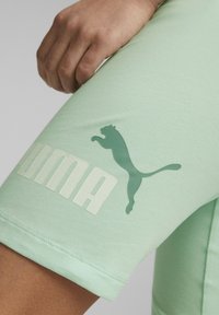 Mátově zelené sportovní šortky vyrobené z měkké tkaniny, se širokým bílým nápisem "PUMA" a tmavě zeleným skákajícím logem pumy.