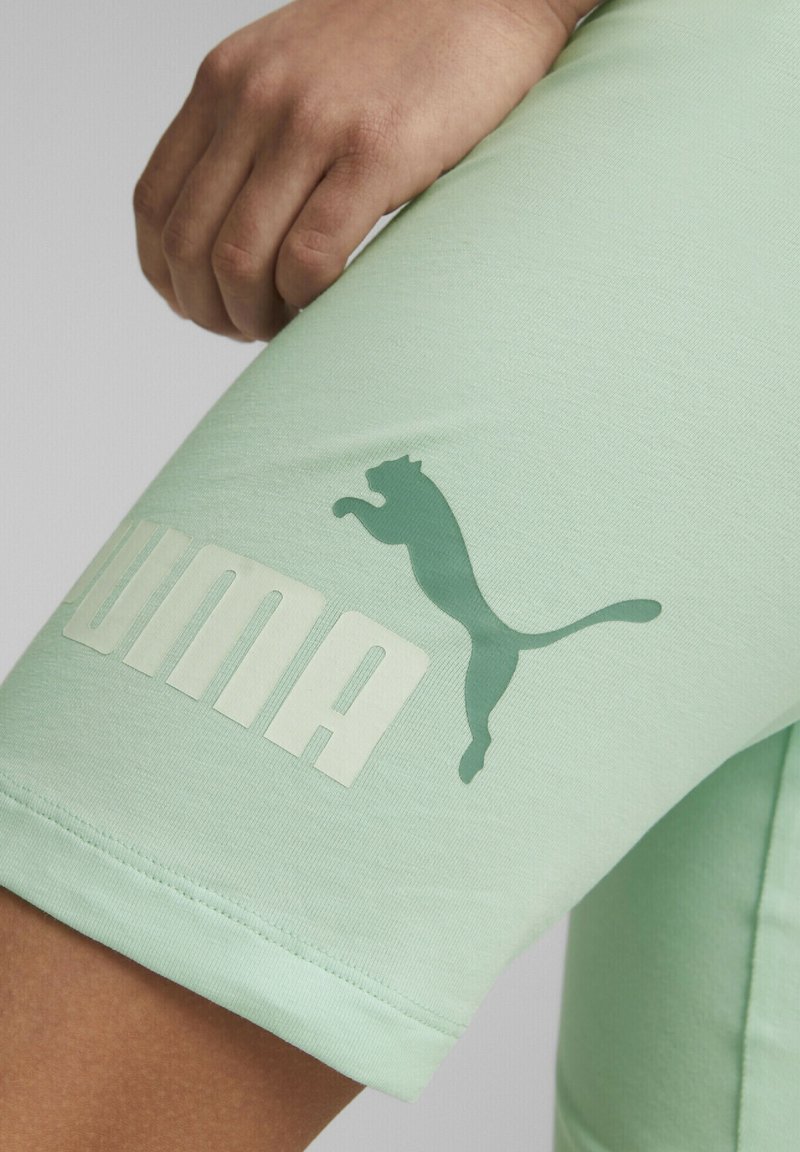 Mátově zelené sportovní šortky vyrobené z měkké tkaniny, se širokým bílým nápisem "PUMA" a tmavě zeleným skákajícím logem pumy.