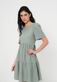 Jasnozielona sukienka z krótkimi bufkami, ozdobiona wzorem w jodełkę i wycięciami typu ażurowego. Rozkloszowany, warstwowy fason z dopasowanym gorsetem.
