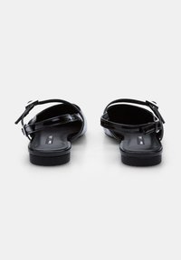 Sandales noires en cuir verni avec une semelle plate, une bride de cheville et une fermeture à boucle. Le design à bout ouvert révèle un intérieur lisse.